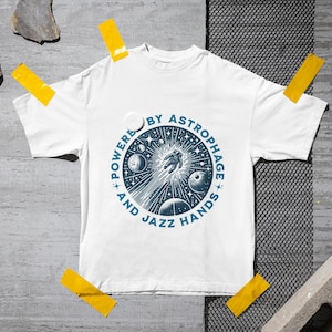 Może przedstawiać: Biała koszulka z niebieską okrągłą grafiką przedstawiającą astronautę, planety i gwiazdy. Tekst "POWERED BY ASTROPHAGE AND JAZZ HANDS" jest wydrukowany wok&oacute;ł grafiki. Koszulka jest przyklejona do szarej powierzchni.