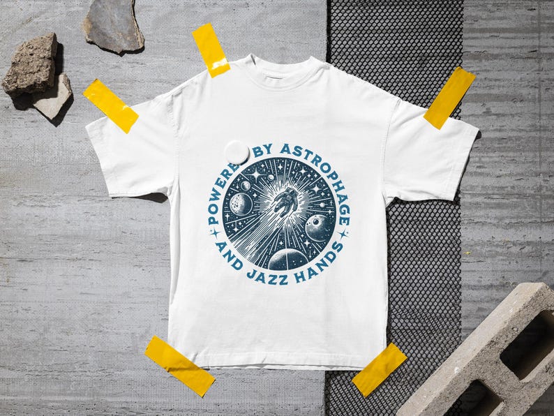 Może przedstawiać: Biała koszulka z niebieską okrągłą grafiką przedstawiającą astronautę, planety i gwiazdy. Tekst "POWERED BY ASTROPHAGE AND JAZZ HANDS" jest wydrukowany wok&oacute;ł grafiki. Koszulka jest przyklejona do szarej powierzchni.