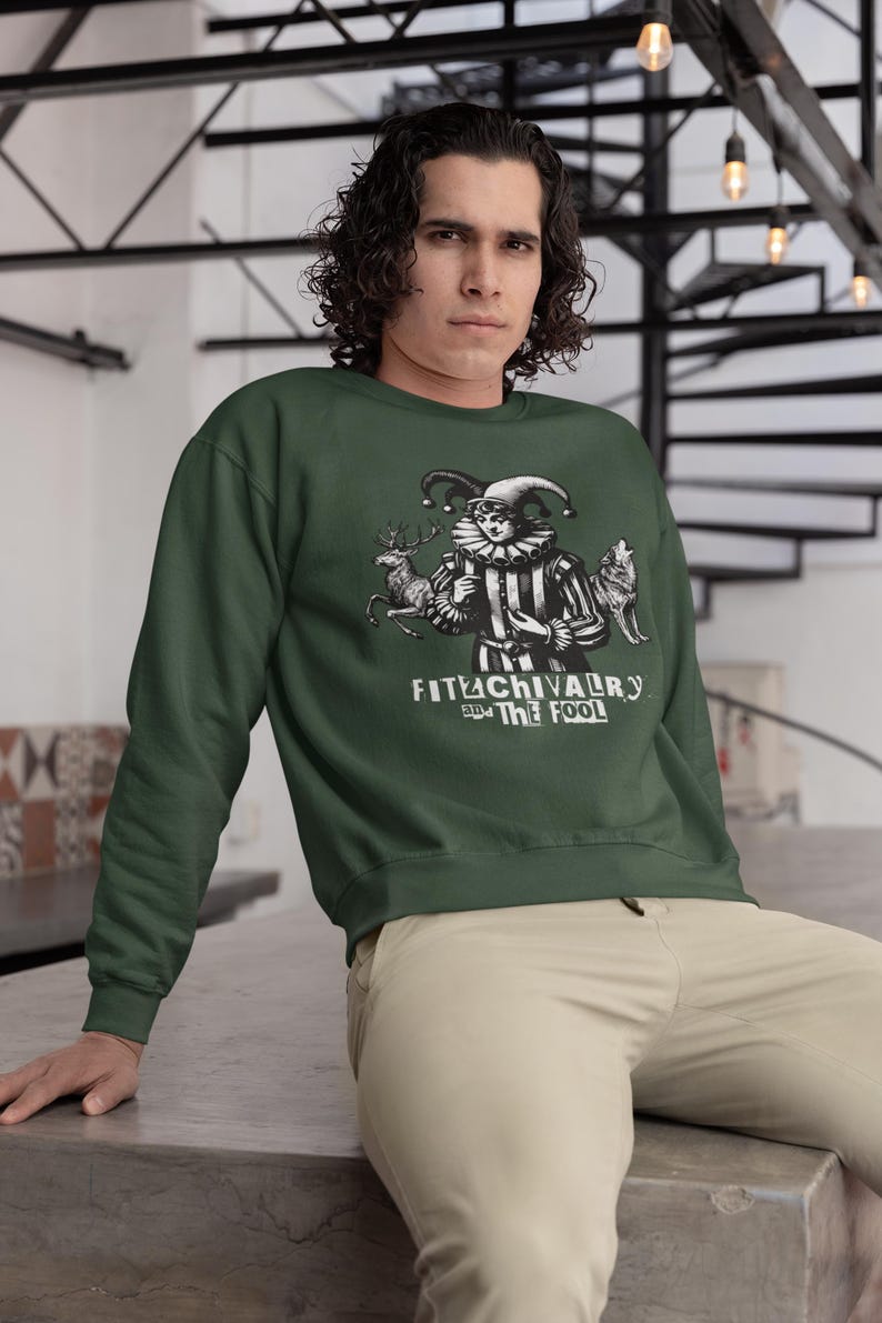 May include: Dark green sweatshirt featuring a black and white illustration of a jester with a deer and wolf. The text "FITACHIVALRY and THE FOOL" is below the illustration. The sweatshirt is a solid color.