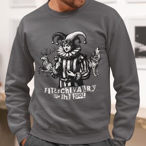 May include: Dark gray sweatshirt featuring a black and white illustration of a jester with a deer and a wolf. The text "FITE CHIVALRY and THE FOOL" is printed below the illustration. The sweatshirt is a crew neck style.