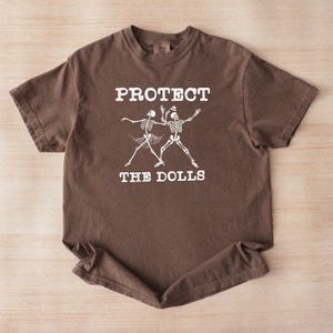 Puede incluir: Camiseta marrón con el texto blanco "PROTECT THE DOLLS" sobre un gráfico de dos esqueletos bailando. La camiseta de manga corta está hecha de un material suave.