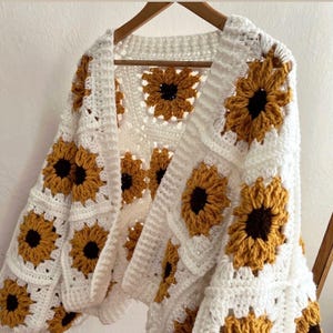 Peut inclure: Un cardigan blanc au crochet avec un motif de tournesol. Le pull ouvert présente des panneaux carrés avec des tournesols jaunes et bruns. Le cardigan est suspendu à un cintre en bois.
