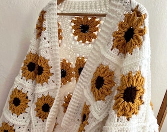 May include: A white crochet cardigan featuring a sunflower design. The open-front sweater has square panels with yellow and brown sunflowers. The cardigan is hanging on a wooden hanger.