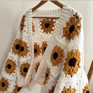 Op de afbeelding: Een witte gehaakte cardigan met een zonnebloemmotief. De cardigan is gemaakt van wit garen, met vierkante panelen van zonnebloemen in geel, bruin en zwart. Het kledingstuk wordt op een houten hanger getoond.