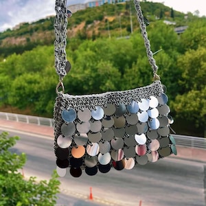 May include: A silver-coloured, rectangular handbag with a chain strap. The bag is covered in overlapping, circular metallic sequins. The chain strap is also silver and made of a woven design. The background shows a green hillside and a road.
