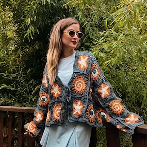 May include: A gray crochet cardigan with orange and white sun and star motifs. The cardigan has a V-neck and long sleeves. The person is wearing a white t-shirt and a light blue skirt. The background is a lush green bamboo forest.