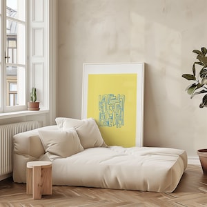 May include: A contemporary living room with a cream chaise longue and two matching cushions. A wooden side table is positioned nearby. A framed abstract art print with a yellow background and blue lines leans against the wall. A potted plant and window are also visible.