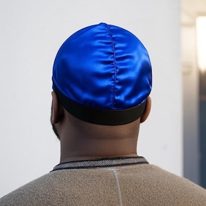 May include: A royal blue satin durag with a black elastic band. The durag is worn on the head and covers the hair. The image is taken from behind, showing the back of the head and neck.