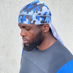 Puede incluir: Un durag de satén azul y negro con estampado de camuflaje y una cola larga. El durag está atado en la cabeza de una persona.