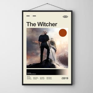 May include: A framed poster for "The Witcher" series. The poster features a central image of two figures on a rocky outcrop, with the title at the top. The color palette includes beige, black, and shades of brown and orange. The year 2019 is printed at the bottom.