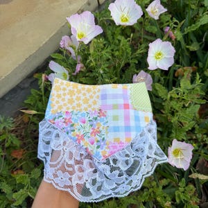 May include: A patchwork fabric skirt with a white lace trim. The skirt features various floral, gingham, and polka dot patterns in pastel colors. The skirt is surrounded by green foliage and pink and white flowers.