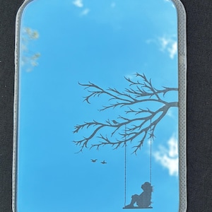 Op de afbeelding: Rechthoekige handspiegel met een blauwe luchtreflectie. Een silhouet van een boomtak met een schommel en een persoon die erop zit, is op de spiegel geëtst. Twee vogels vliegen.