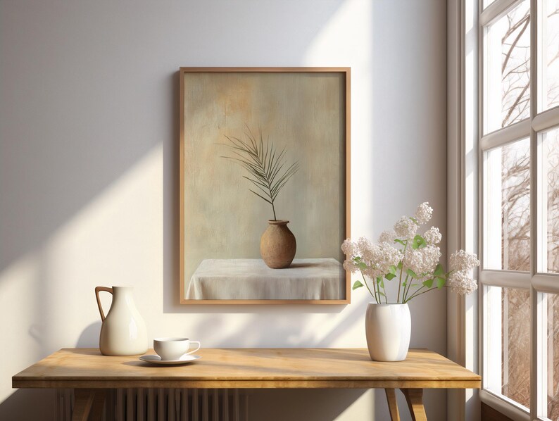 May include: A framed painting of a single brown vase with a leafy branch on a white tablecloth. The painting is hanging on a white wall with a wooden frame.