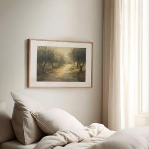 Puede incluir: Una pintura de paisaje enmarcada representa un camino soleado bordeado de olivos. La obra de arte, en tonos c&aacute;lidos de marr&oacute;n, verde y amarillo, est&aacute; enmarcada en un marco de madera clara. La pintura cuelga en una pared de color neutro sobre una cama con ropa de cama color crema.