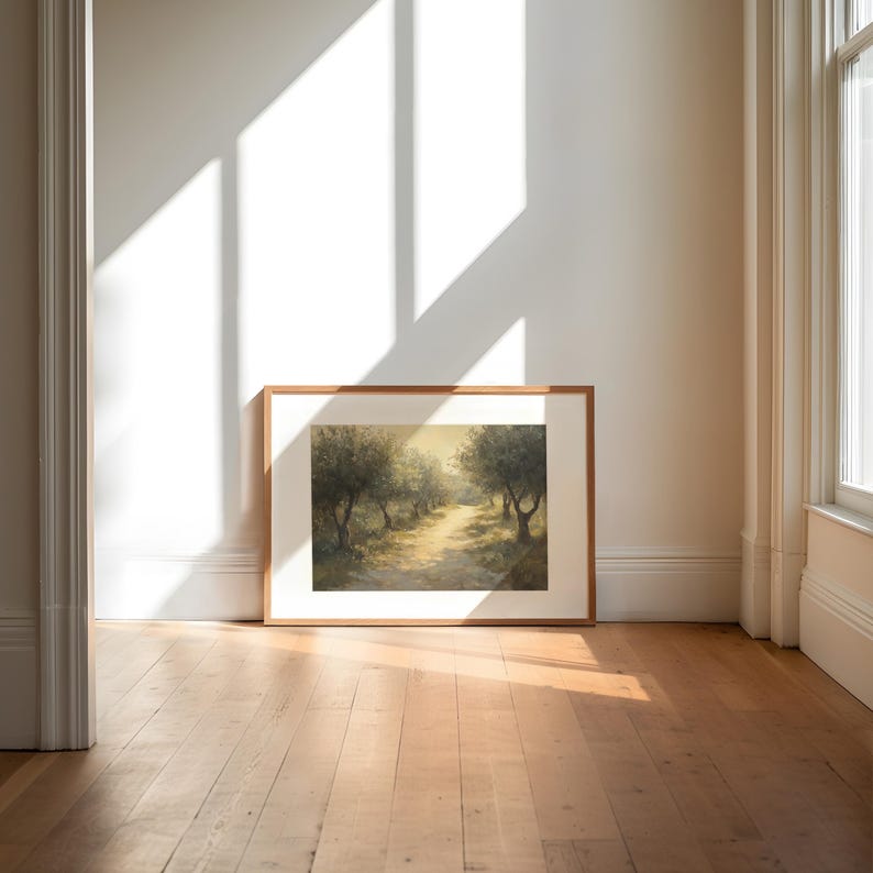 Puede incluir: Una pintura enmarcada de un paisaje con un camino bordeado de &aacute;rboles, ba&ntilde;ado por el sol. La obra de arte, en un marco de madera, se apoya en una pared blanca, con luz natural de una ventana. El suelo es de madera clara.