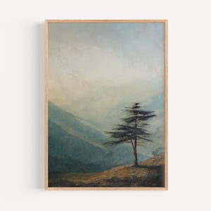May include: A framed landscape art print featuring a solitary tree on a hillside. The artwork uses muted blues, greens, and browns to depict a misty mountain scene. The print is encased in a light-colored wooden frame.