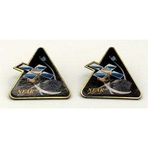 May include: Two triangular metal pins featuring a space theme. Each pin displays a gold-colored border, a black background, and an illustration of the NEAR spacecraft orbiting a gray asteroid. The text NEAR is printed in gold at the bottom left.