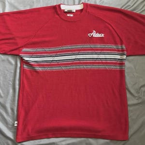 May include: A red short-sleeve knit shirt featuring a horizontal chest band with a white, black, and grey striped pattern overlaid with a diamond stitch design. The word Avirex is embroidered in white on the upper left chest area of the shirt.