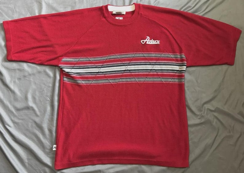 May include: A red short-sleeve knit shirt featuring a horizontal chest band with a white, black, and grey striped pattern overlaid with a diamond stitch design. The word Avirex is embroidered in white on the upper left chest area of the shirt.