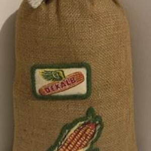 May include: A burlap sack with a drawstring closure. The sack features two embroidered patches: one with the word "DEKALB" and an ear of corn, and another of an ear of corn. The sack is a natural tan color.