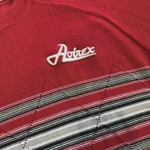 May include: A red knit sweater featuring the white embroidered text Avirex on the upper chest. The lower portion of the garment displays a horizontal band with a repeating diamond pattern in shades of red, white, gray, and black stripes.