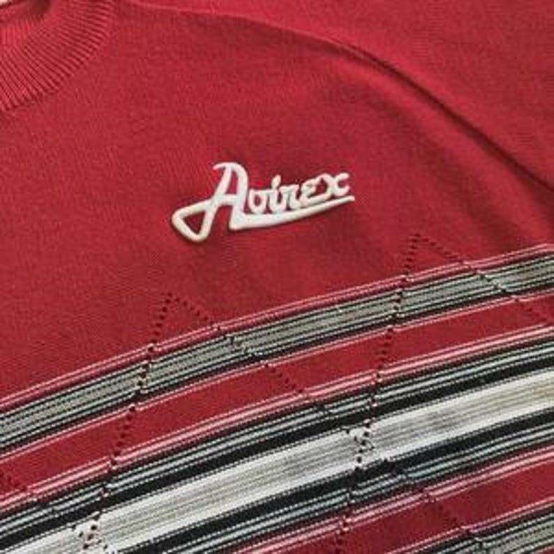 May include: A red knit sweater featuring the white embroidered text Avirex on the upper chest. The lower portion of the garment displays a horizontal band with a repeating diamond pattern in shades of red, white, gray, and black stripes.