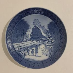 May include: A blue and white decorative plate featuring a winter scene with a train, trees, and figures. The plate has a textured border and the year "1973" at the bottom. The plate is round.