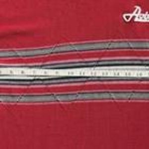 May include: A red long-sleeve shirt featuring the white text Avirex on the upper chest and a horizontal band with grey, black, and white stripes across the midsection. A white measuring tape shows a width of approximately 23 inches across.