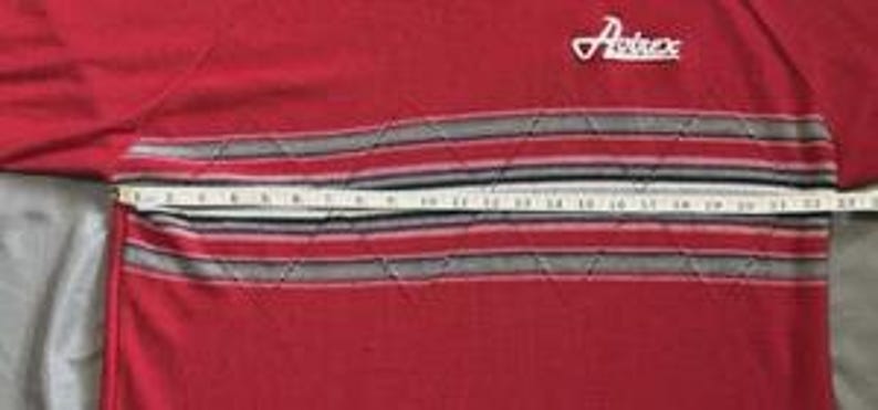 May include: A red long-sleeve shirt featuring the white text Avirex on the upper chest and a horizontal band with grey, black, and white stripes across the midsection. A white measuring tape shows a width of approximately 23 inches across.