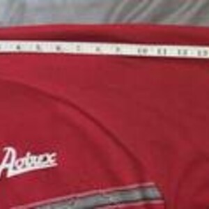 May include: A red t-shirt with a white collar and the text Avirex embroidered on the chest. A white measuring tape is placed across the sleeve, showing a length of approximately 20 inches from the collar seam to the edge of the sleeve.