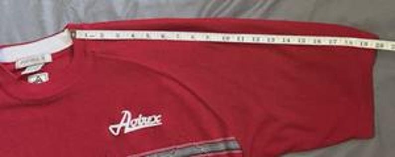 May include: A red t-shirt with a white collar and the text Avirex embroidered on the chest. A white measuring tape is placed across the sleeve, showing a length of approximately 20 inches from the collar seam to the edge of the sleeve.