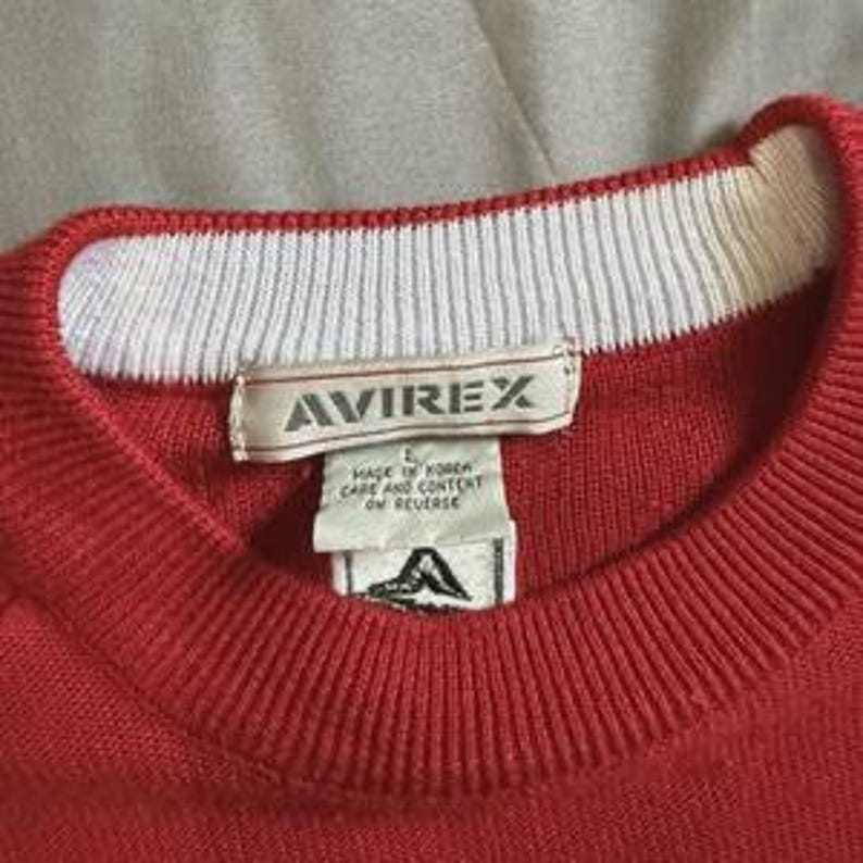 May include: A red knit sweater featuring a white ribbed crew neckline. A white fabric label is stitched inside the collar with the text AVIREX, followed by a smaller tag stating Made in Korea, care and content on reverse, and a logo graphic.