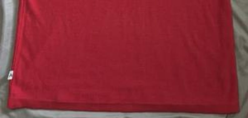 May include: A solid red t-shirt featuring a small white rectangular tag attached to the lower left side seam. The fabric has a visible knit texture and a consistent, vibrant red color throughout the entire garment, ending in a straight hem.
