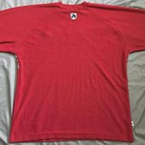 May include: A solid red short-sleeve t-shirt featuring a small white and black shield-shaped logo patch centered just below the crew neckline. The garment has a simple, plain design with a small white rectangular tag visible on the lower side seam.