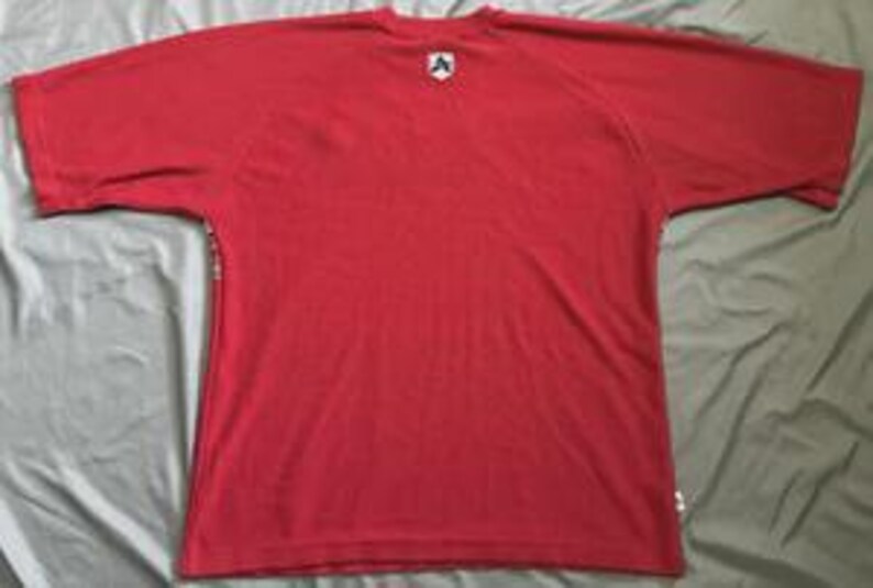 May include: A solid red short-sleeve t-shirt featuring a small white and black shield-shaped logo patch centered just below the crew neckline. The garment has a simple, plain design with a small white rectangular tag visible on the lower side seam.