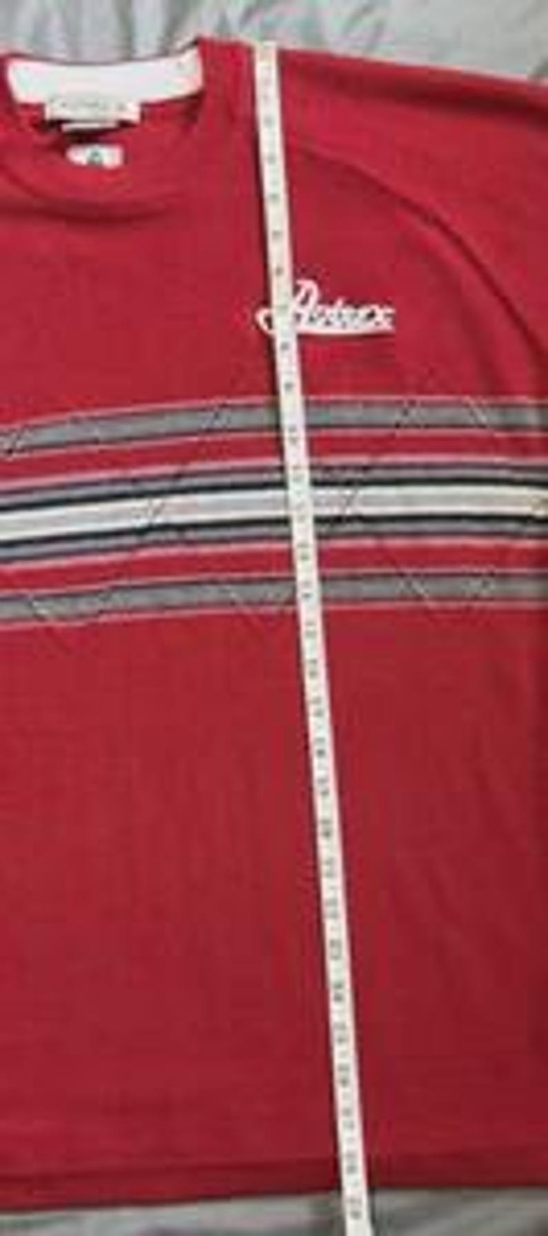 May include: A red long-sleeve shirt featuring a white Artex logo on the chest and a horizontal band with grey, black, and white stripes. A white measuring tape is placed vertically over the shirt, showing a length of approximately 29 inches.