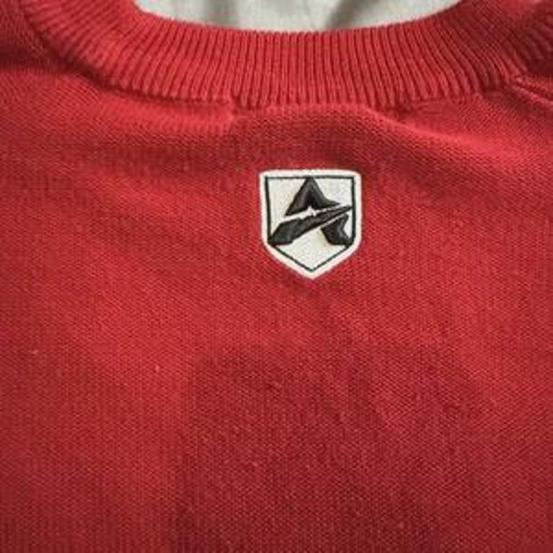 May include: A bright red knit sweater featuring a ribbed crew neckline. Centered on the chest is a white shield-shaped patch with a black embroidered logo that depicts a stylized letter A with a horizontal line through the center of the character.