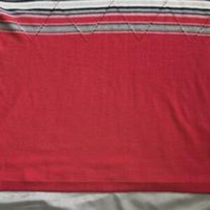 May include: A red knit shirt featuring a horizontal striped pattern across the upper chest in shades of white, black, and grey. The striped section includes a subtle diamond-shaped stitched texture. A small white brand tag is visible on the lower left side.