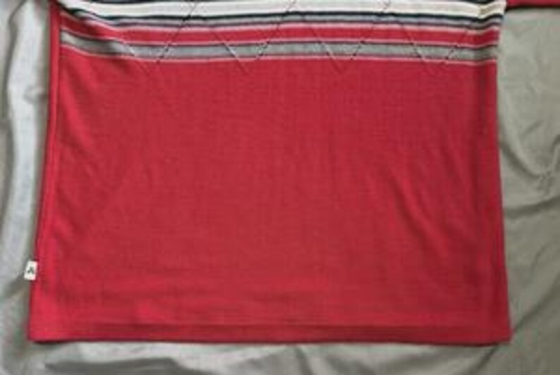 May include: A red knit shirt featuring a horizontal striped pattern across the upper chest in shades of white, black, and grey. The striped section includes a subtle diamond-shaped stitched texture. A small white brand tag is visible on the lower left side.