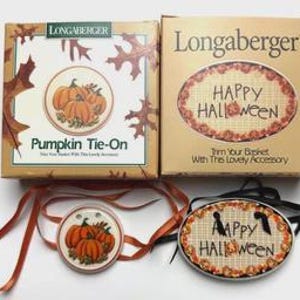 May include: Two Longaberger boxes and two tie-on basket accessories. One box reads "Pumpkin Tie-On" with a pumpkin graphic. The other box reads "Happy Halloween." The accessories feature pumpkin and Halloween designs.