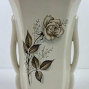 May include: A cream-colored ceramic vase with a flared top and two side handles. The front features a detailed illustration of a single rose with leaves and small sprigs in shades of brown and gray, centered on the smooth, glossy surface.
