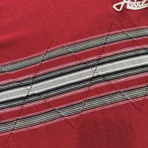 May include: A red knit sweater featuring a horizontal band with alternating stripes in black, white, and grey. The band is overlaid with a stitched diamond pattern. A white embroidered logo reading Avirex is visible in the upper right corner.