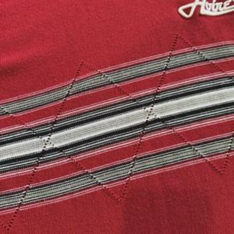 May include: A red knit sweater featuring a horizontal band with alternating stripes in black, white, and grey. The band is overlaid with a stitched diamond pattern. A white embroidered logo reading Avirex is visible in the upper right corner.