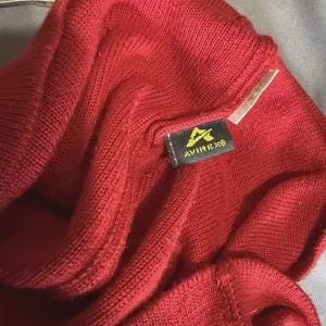May include: A bright red ribbed knit beanie featuring a black rectangular fabric tag sewn onto the exterior. The tag displays a yellow graphic logo above the text AVIREX in white capital letters. The knit texture is consistent throughout.