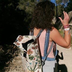 Puede incluir: Mochila crema con estampado floral, cierre enrollable y hebilla negra. La mochila presenta un patrón de flores y hojas en tonos rosas, marrones y verdes. Correas grises e interior negro completan el diseño.
