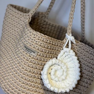 May include: Beige crochet tote bag with matching handles and a white spiral-shaped charm. The bag features a woven texture and a small label that reads "A.S. Knitting Accessories".