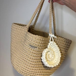 Peut inclure: Un sac fourre-tout beige en crochet avec des anses tressées et un charm en forme de spirale blanche. Le sac a une forme arrondie et une étiquette qui dit "AS.KNITTING accessories". Le sac est tenu par une personne.