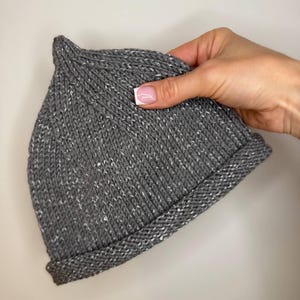 May include: A gray knit beanie with a folded brim. The hat has a speckled pattern with white flecks throughout the gray yarn. The beanie is a classic style, suitable for cold weather.