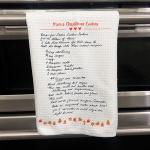 May include: A white kitchen towel with a handwritten recipe for "Mom's Christmas Cookies". The recipe includes ingredients like shortening, sugar, milk, vanilla, baking powder, and flour. The towel has a red border and a festive design with small red hearts and orange Christmas ornaments.