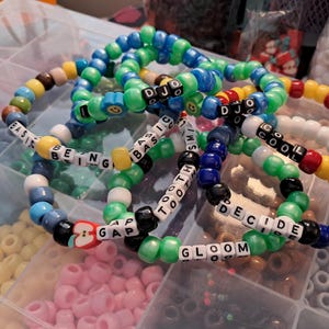 May include: A collection of colorful beaded bracelets. Some bracelets feature white letter beads spelling out words like "COOL", "GLOOM", and "DECIDE". The bracelets are in shades of blue, green, yellow, and red.